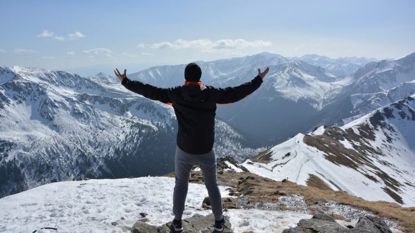 A man stands on a snowy mountain peak with arms raised, embracing the breathtaking view.
