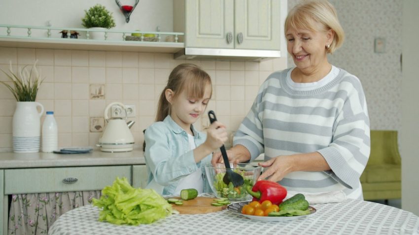 Little girl is cooking salad mixing vegetables in bowl and chatting to cheerful granny in kitchen at home. Modern lifestyle, meals preparation and family concept.